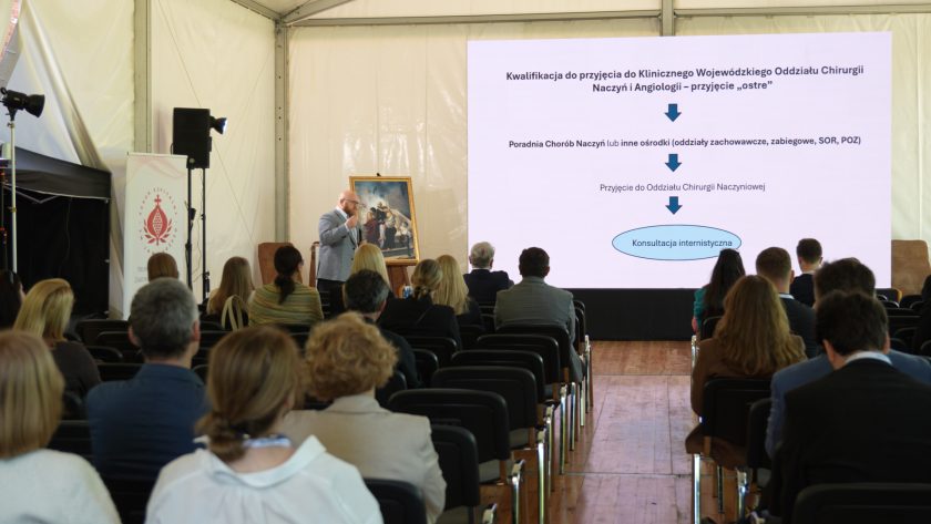 Prelegent prowadzi wykład w dużym namiocie konferencyjnym. Na ekranie wyświetlony jest schemat dotyczący kwalifikacji pacjentów do przyjęcia do oddziału chirurgii naczyń i angiologii. Publiczność siedzi w rzędach krzeseł i słucha wystąpienia