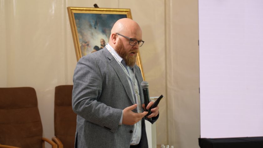 Mężczyzna z brodą w szarej marynarce przemawia do mikrofonu podczas wykładu. W dłoni trzyma pilot do prezentacji. Za nim widoczny jest obraz na sztaludze oraz fragment dużego ekranu prezentacyjnego