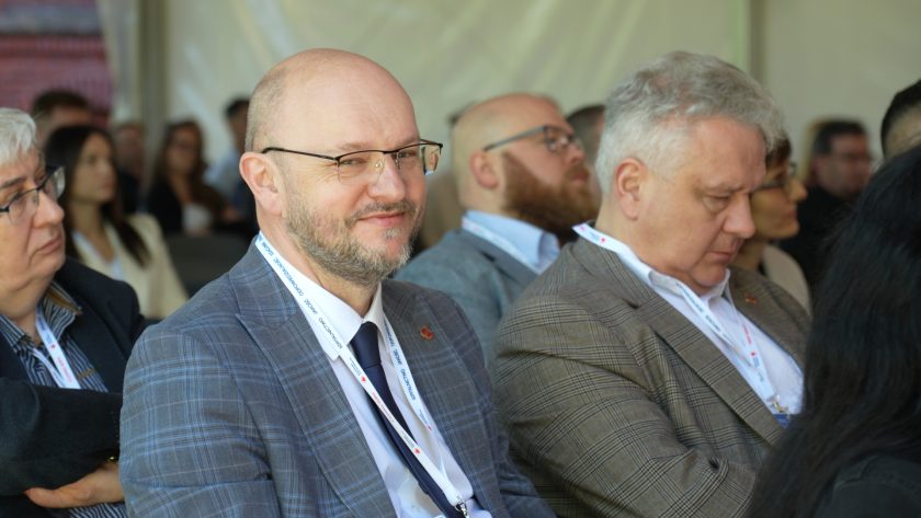 Uczestnik konferencji w jasnoszarej marynarce siedzi wśród publiczności i lekko uśmiecha się w stronę aparatu. Obok niego siedzą inni goście wydarzenia z identyfikatorami konferencyjnymi
