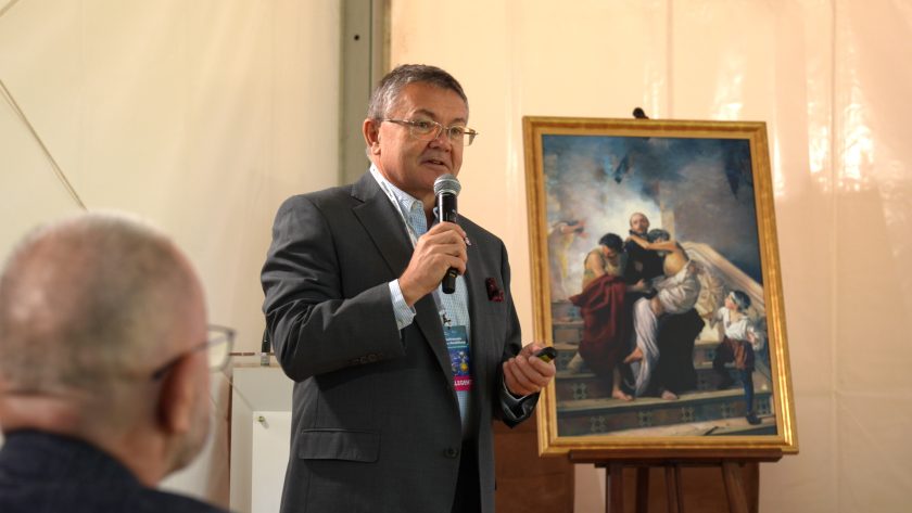 Prelegent w ciemnym garniturze przemawia do mikrofonu podczas konferencji medycznej. W tle znajduje się obraz na sztaludze, a na pierwszym planie widać fragment głowy jednego z uczestników spotkania