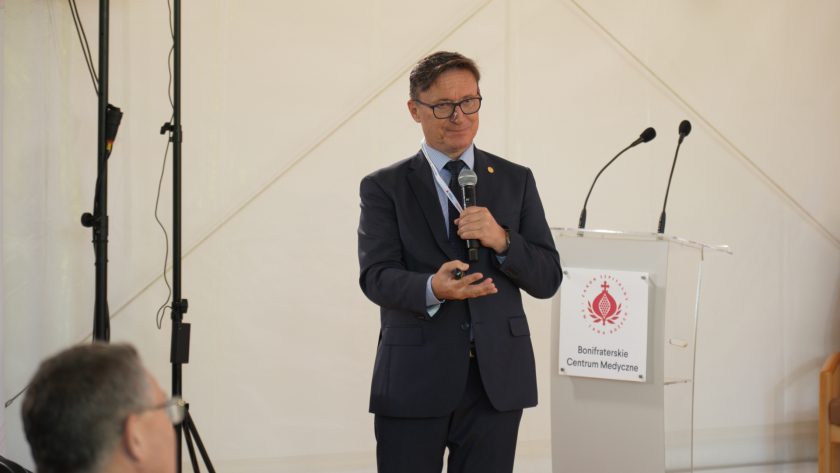 Prelegent w ciemnym garniturze stoi przy pulpicie Bonifraterskiego Centrum Medycznego i przemawia do mikrofonu. Wystąpienie odbywa się w namiocie konferencyjnym, a mężczyzna trzyma w dłoni pilot do prezentacji