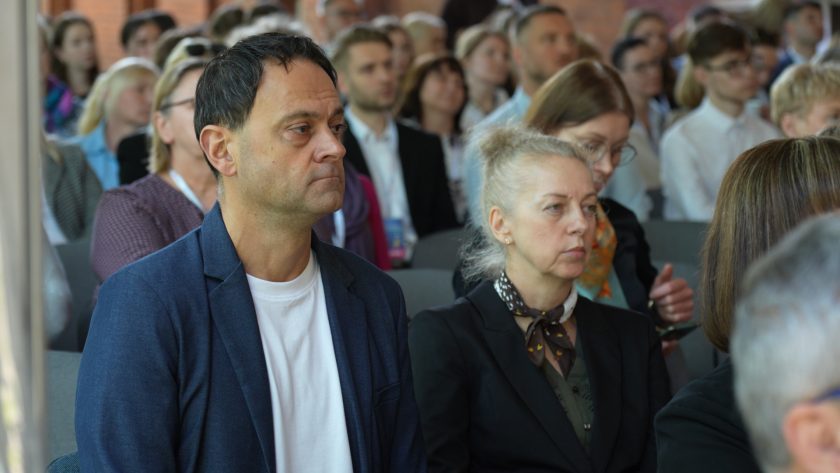 Uczestnicy konferencji siedzą w rzędach krzeseł i słuchają wystąpienia. Na pierwszym planie widoczni są kobieta i mężczyzna w eleganckich strojach, skupieni na przebiegu prelekcji. W tle znajduje się licznie zgromadzona publiczność
