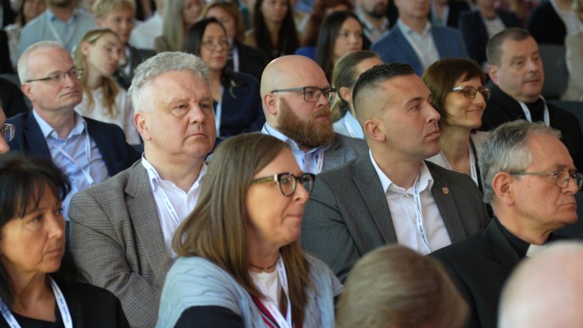 Publiczność zgromadzona w namiocie konferencyjnym słucha prelekcji. W centralnej części kadru widoczni są uczestnicy siedzący blisko siebie, skupieni na wystąpieniu odbywającym się poza kadrem