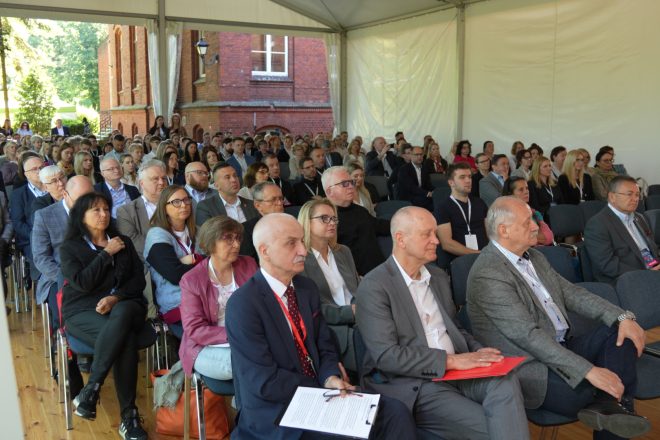 Szerokie ujęcie sali konferencyjnej w dużym namiocie. Licznie zgromadzona publiczność siedzi w rzędach krzeseł, a przez otwarte ściany namiotu widać ceglany budynek i otoczenie zieleni