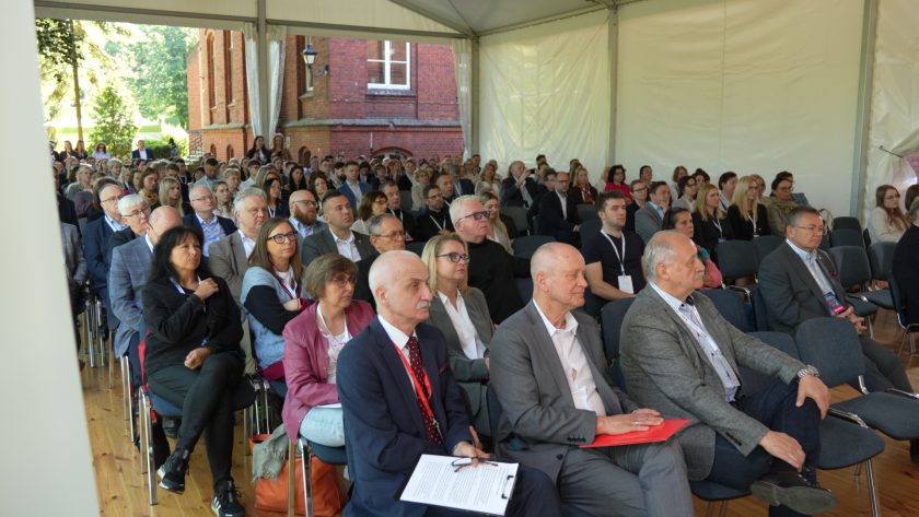 Szerokie ujęcie sali konferencyjnej w dużym namiocie. Licznie zgromadzona publiczność siedzi w rzędach krzeseł, a przez otwarte ściany namiotu widać ceglany budynek i otoczenie zieleni