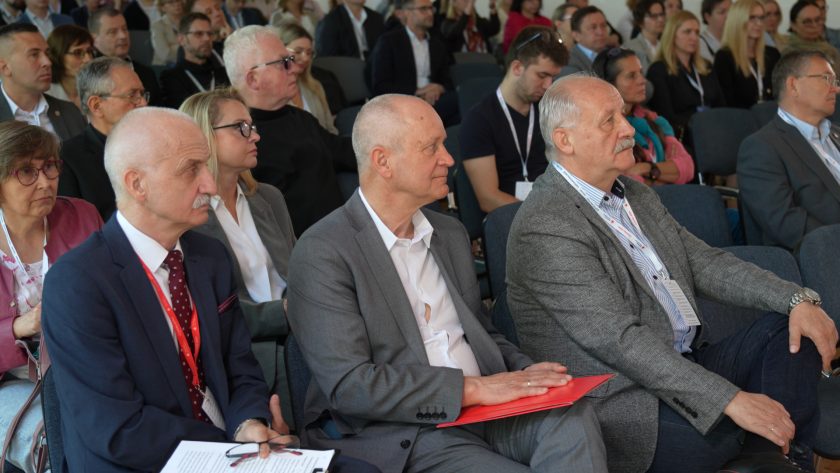 Uczestnicy konferencji siedzą w pierwszych rzędach i słuchają wykładu. Na pierwszym planie widoczni są mężczyźni w garniturach, jeden z nich trzyma czerwony folder z dokumentami