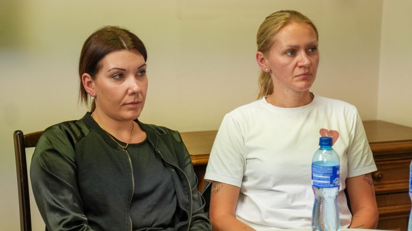 Dwie kobiety siedzące przy stole uczestniczą w dyskusji podczas spotkania. Kobieta po lewej stronie ubrana jest w czarną kurtkę i ciemną bluzkę, natomiast kobieta po prawej ma biały T-shirt z nadrukiem serca. Obie z powagą i skupieniem słuchają wypowiedzi prowadzącego. Na pierwszym planie widoczna jest butelka wody stojąca na stole