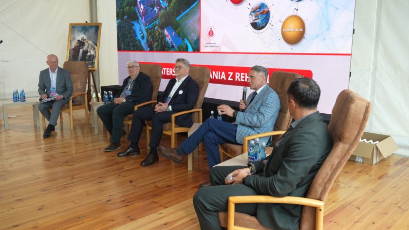 Panel dyskusyjny podczas konferencji z udziałem kilku prelegentów siedzących w fotelach na scenie. Jeden z mężczyzn przemawia do mikrofonu, a pozostali uczestnicy słuchają jego wypowiedzi. W tle wyświetlana jest prezentacja związana z rehabilitacją i działalnością Bonifraterskiego Centrum Medycznego