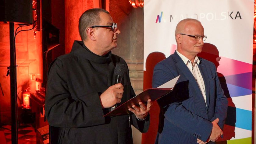 Przedstawiciel Zakonu Bonifratrów przemawia do zgromadzonej publiczności, trzymając mikrofon i okolicznościową teczkę. Obok stoi przedstawiciel Województwa Małopolskiego, partnera wydarzenia
