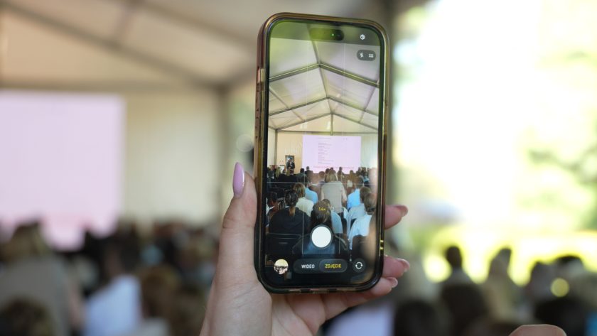 Telefon komórkowy trzymany w dłoni rejestruje wykład odbywający się w namiocie konferencyjnym. Na ekranie telefonu widać prelegenta, publiczność i duży ekran prezentacyjny