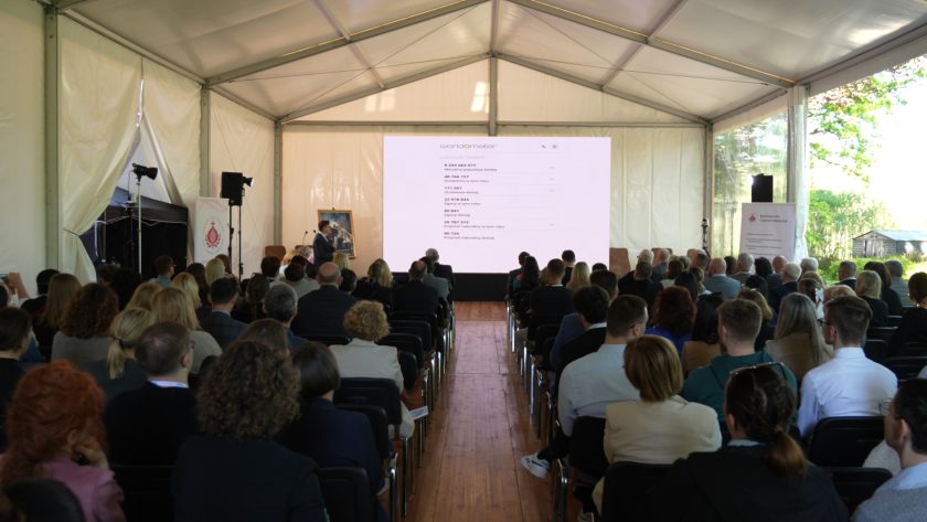 Szerokie ujęcie wykładu w namiocie konferencyjnym. Prelegent stoi przy dużym ekranie, na którym wyświetlana jest prezentacja, a publiczność siedzi w równych rzędach i obserwuje wystąpienie