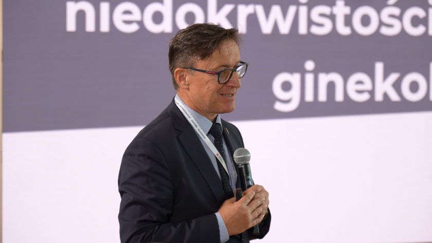 Prelegent w ciemnym garniturze przemawia do mikrofonu na tle dużego ekranu prezentacyjnego. W tle widoczny jest fragment slajdu dotyczącego niedokrwistości i zagadnień ginekologicznych