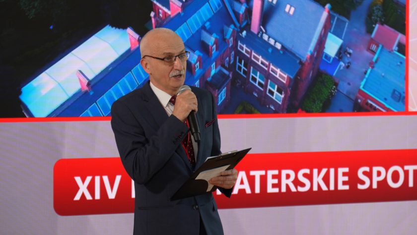 Mężczyzna w garniturze prowadzi wystąpienie przed dużym ekranem z grafiką wydarzenia. Trzyma mikrofon oraz notatki na podkładce, a za nim widoczny jest napis odnoszący się do XIV Bonifraterskich Spotkań z Rehabilitacją
