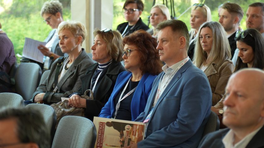 Uczestnicy konferencji siedzą w rzędach krzeseł i słuchają prelekcji. Na pierwszym planie kobiety i mężczyzna z identyfikatorami konferencyjnymi skupiają uwagę na wystąpieniu, a jedna z osób trzyma gazetę „Bonifratrzy w służbie chorym”