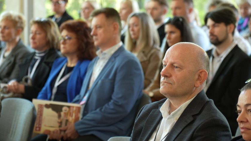 Uczestnicy konferencji siedzą w rzędach krzeseł i z uwagą słuchają wystąpienia. Na pierwszym planie widoczny jest mężczyzna w ciemnej marynarce i białej koszuli, a za nim pozostali goście wydarzenia z identyfikatorami konferencyjnymi