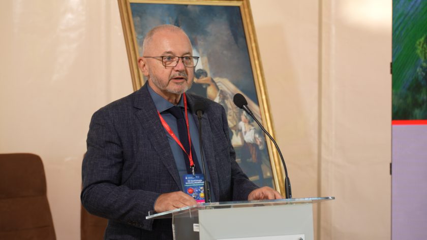 Mężczyzna w granatowej marynarce stoi za mównicą Bonifraterskiego Centrum Medycznego i przemawia do mikrofonów. Za nim znajduje się obraz w złotej ramie oraz fragment dużego ekranu prezentacyjnego. Prelegent ma na szyi identyfikator wydarzenia