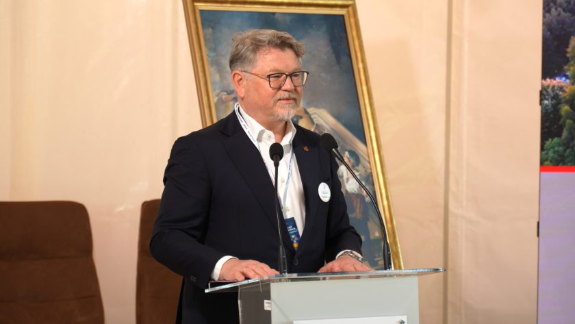 Mężczyzna w ciemnym garniturze stoi za mównicą i przemawia podczas części oficjalnej konferencji. W tle znajduje się obraz na sztaludze oraz fragment ekranu prezentacyjnego. Na mównicy widoczne jest logo Bonifraterskiego Centrum Medycznego