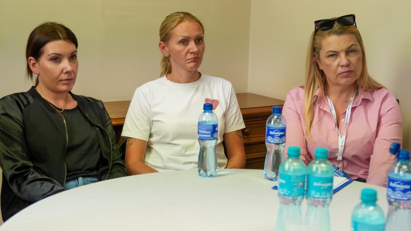 Trzy uczestniczki spotkania siedzą przy stole i słuchają rozmowy. Po lewej stronie znajduje się kobieta w czarnej kurtce, obok niej kobieta w białym T-shircie, a po prawej blondynka w różowej koszuli z okularami przeciwsłonecznymi opartymi na głowie. Na stole ustawione są butelki wody mineralnej. Wszystkie kobiety mają skupione i uważne wyrazy twarzy