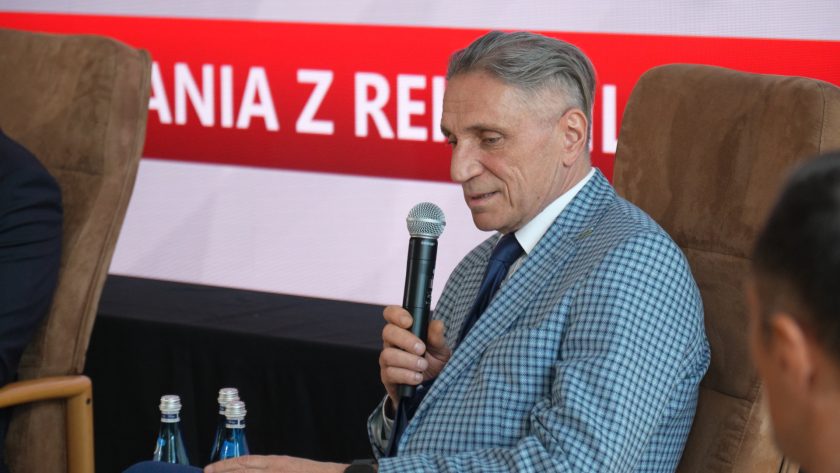 Mężczyzna w eleganckiej marynarce w kratę siedzi na scenie konferencyjnej i przemawia do mikrofonu podczas panelu dyskusyjnego. Za nim widoczny jest ekran z czerwonym napisem odnoszącym się do Spotkań z Rehabilitacją