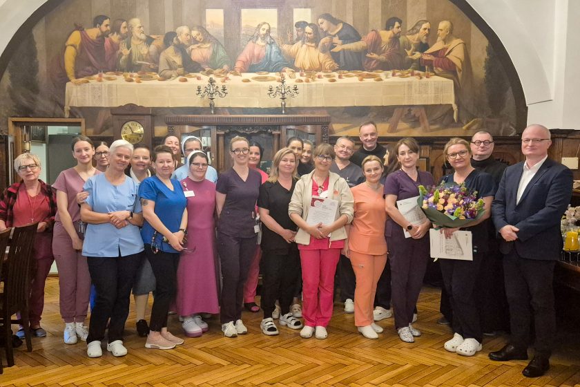 Grupowe zdjęcie pracowników szpitala, przedstawicieli dyrekcji i zakonników w historycznej sali z malowidłem przedstawiającym Ostatnią Wieczerzę