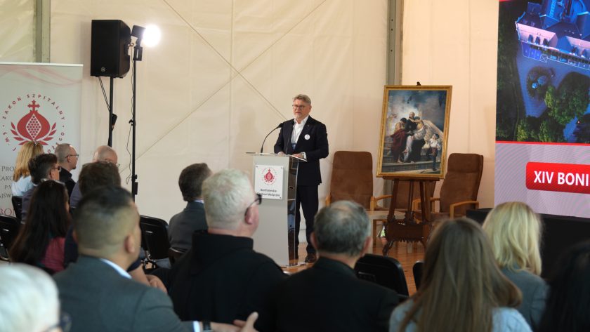 Prelegent przemawia do uczestników konferencji z mównicy ustawionej obok dużego ekranu i obrazu na sztaludze. Na pierwszym planie widać siedzących słuchaczy, a z boku baner z symbolem Zakonu Szpitalnego św. Jana Bożego