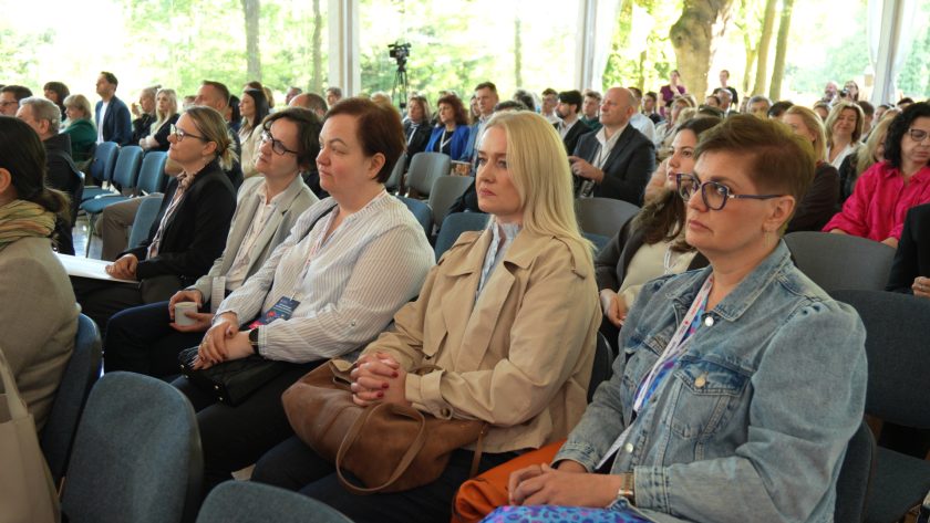 Uczestniczki i uczestnicy konferencji siedzą w rzędach krzeseł i słuchają wykładu. Na pierwszym planie widoczne są kobiety z identyfikatorami, skupione na prezentacji. W tle widać licznie zgromadzoną publiczność oraz jasne wnętrze namiotu