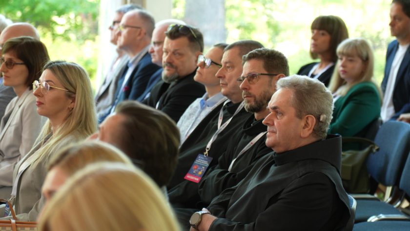 Rząd uczestników konferencji, w tym osoby duchowne w czarnych habitach, słucha wystąpienia. Ujęcie z boku pokazuje skupione twarze słuchaczy oraz identyfikatory zawieszone na smyczach. W tle widoczni są kolejni goście wydarzenia
