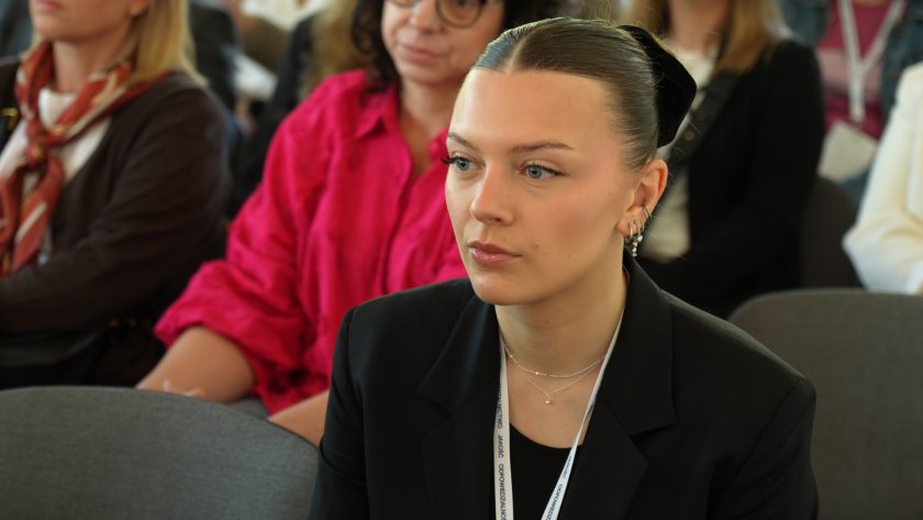 Zbliżenie na młodą uczestniczkę konferencji siedzącą wśród publiczności. Kobieta w czarnej marynarce i identyfikatorze patrzy w stronę sceny. Za nią widoczne są inne osoby uczestniczące w wykładzie