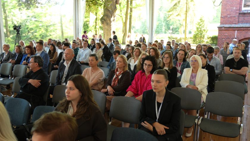 Szerokie ujęcie publiczności siedzącej w namiocie konferencyjnym podczas XIV Bonifraterskich Spotkań z Rehabilitacją. Uczestnicy zajmują miejsca w rzędach krzeseł i z uwagą słuchają wystąpienia. W tle, przez przezroczyste ściany namiotu, widoczne są drzewa oraz fragment ceglanego budynku