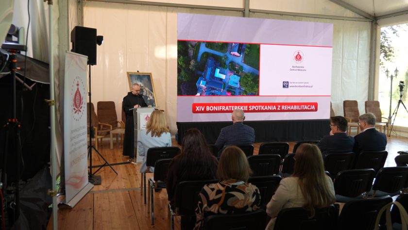 Prelegent występuje przy mównicy podczas XIV Bonifraterskich Spotkań z Rehabilitacją. Na dużym ekranie za nim widoczna jest grafika wydarzenia z napisem „Prehabilitacja jako nieodzowny element leczenia planowego” oraz zdjęciem kompleksu z lotu ptaka. Przed sceną siedzą uczestnicy konferencji