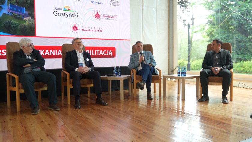 Szerokie ujęcie panelu konferencyjnego z udziałem czterech prelegentów siedzących na scenie w fotelach. Jeden z uczestników przemawia do mikrofonu, a za nimi widoczny jest duży ekran z logotypami partnerów wydarzenia oraz informacjami o Bonifraterskich Spotkaniach z Rehabilitacją