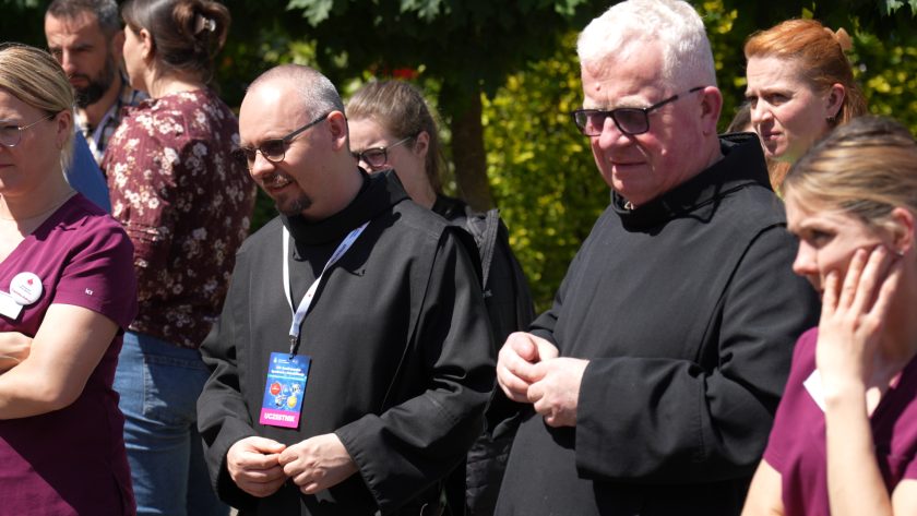 Grupa uczestników wydarzenia stoi na świeżym powietrzu w słoneczny dzień. W centrum kadru widoczni są dwaj zakonnicy w czarnych habitach i identyfikatorach konferencyjnych. Wokół nich stoją uczestnicy spotkania, obserwujący pokaz lub prezentację odbywającą się poza kadrem