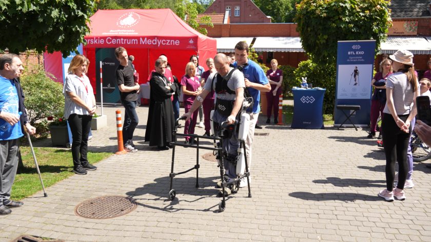 Na dziedzińcu Bonifraterskiego Centrum Medycznego odbywa się pokaz nowoczesnego sprzętu rehabilitacyjnego. Mężczyzna korzysta z egzoszkieletu wspomagającego chodzenie, asekurowany przez instruktora. Wokół zgromadzili się uczestnicy wydarzenia, a w tle widoczne są namioty promocyjne i banery firm związanych z rehabilitacją