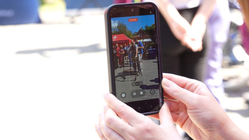 Zbliżenie na telefon komórkowy trzymany w dłoniach uczestniczki wydarzenia. Na ekranie widoczny jest nagrywany pokaz osoby korzystającej z egzoszkieletu rehabilitacyjnego podczas prezentacji na świeżym powietrzu