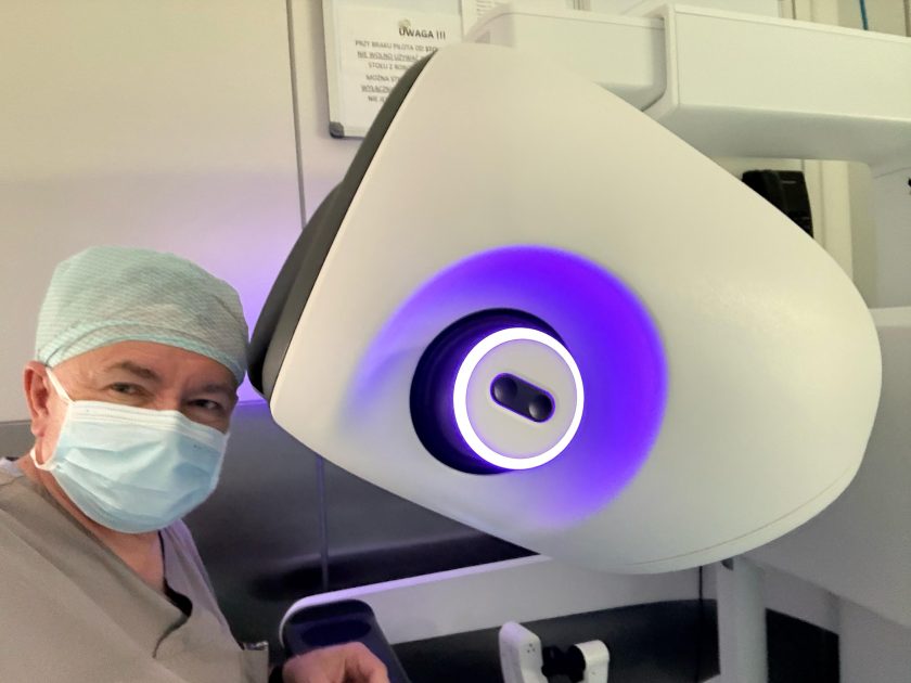 Lekarz ubrany w strój operacyjny, czepek i maseczkę stoi obok konsoli systemu chirurgii robotycznej da Vinci. Na pierwszym planie widoczny jest duży biały wizjer urządzenia z charakterystycznym fioletowym podświetleniem, przeznaczony do obserwacji pola operacyjnego w technologii 3D