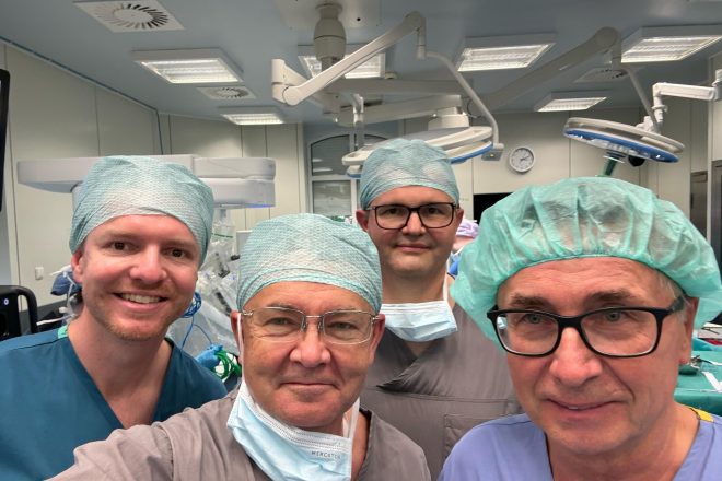 Czterech członków zespołu medycznego pozuje do wspólnego zdjęcia na sali operacyjnej. Lekarze ubrani są w stroje chirurgiczne, czepki i częściowo opuszczone maseczki. W tle widoczne są lampy operacyjne oraz elementy robota chirurgicznego przygotowanego do zabiegu