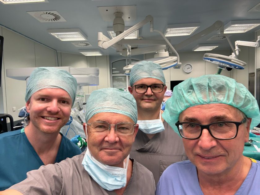 Czterech członków zespołu medycznego pozuje do wspólnego zdjęcia na sali operacyjnej. Lekarze ubrani są w stroje chirurgiczne, czepki i częściowo opuszczone maseczki. W tle widoczne są lampy operacyjne oraz elementy robota chirurgicznego przygotowanego do zabiegu