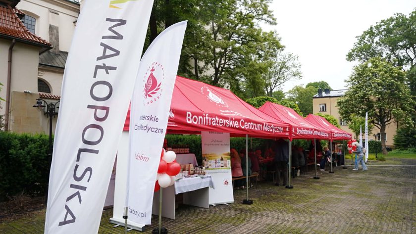 Namioty Bonifraterskiego Centrum Medycznego ustawione na dziedzińcu wśród zieleni. Przed nimi stoją flagi promujące działalność Bonifratrów