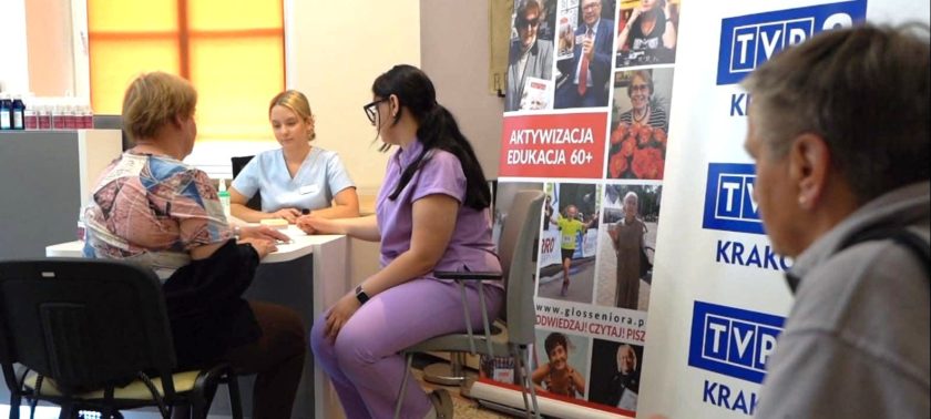 Uczestniczki wydarzenia siedzą przy stoliku konsultacyjnym. W tle widoczne są roll-upy Głosu Seniora i TVP3 Kraków