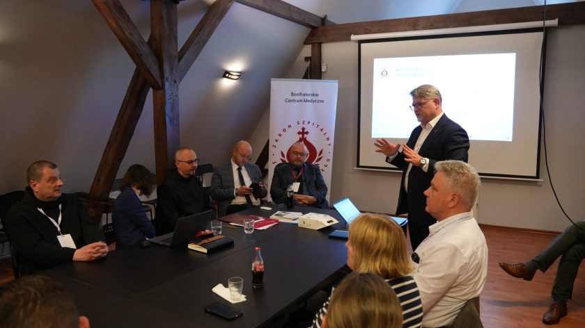 Mężczyzna w granatowym garniturze stoi przy stole konferencyjnym i przemawia do uczestników spotkania. Za nim znajduje się ekran projekcyjny z wyświetloną prezentacją oraz baner Bonifraterskiego Centrum Medycznego. Pozostali uczestnicy siedzą wokół stołu i słuchają wystąpienia, część z nich ma przed sobą laptopy oraz dokumenty