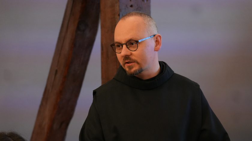 Brat zakonny w czarnym habicie i okularach przemawia podczas spotkania. W tle widoczne są drewniane belki poddasza oraz fragment sali konferencyjnej. Duchowny zwrócony jest w stronę uczestników i wygląda na zaangażowanego w prowadzoną dyskusję