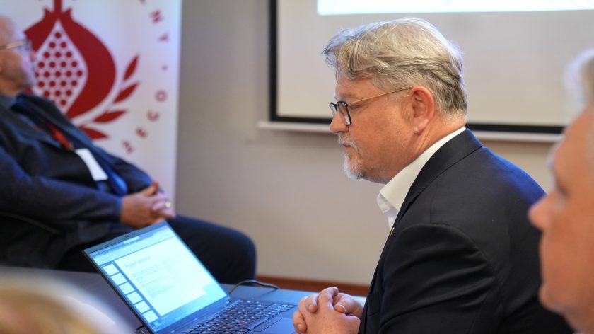 Mężczyzna w okularach i granatowej marynarce siedzi przy stole konferencyjnym obok otwartego laptopa z wyświetloną prezentacją. W tle widać ekran projekcyjny oraz innych uczestników spotkania. Atmosfera skupienia wskazuje na trwającą prezentację lub debatę
