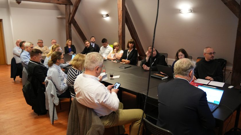Szerokie ujęcie sali konferencyjnej podczas spotkania. Uczestnicy siedzą wokół dużego stołu i słuchają wystąpienia jednego z zakonników. Na stole znajdują się laptopy, telefony, notatniki oraz dokumenty. W pomieszczeniu dominują drewniane elementy konstrukcyjne poddasza