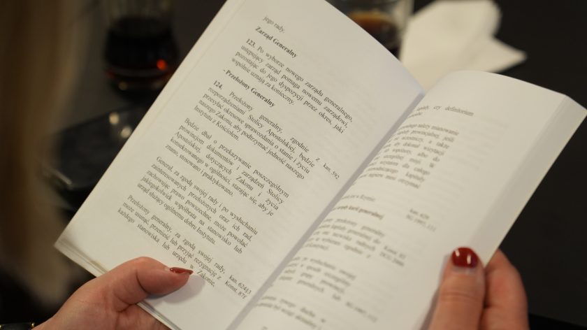 Zbliżenie na otwartą książkę trzymaną w dłoniach uczestniczki spotkania. Na widocznej stronie znajduje się tekst dotyczący zarządu generalnego i przełożonego generalnego. W tle rozmyty fragment stołu konferencyjnego i szklanka z napojem