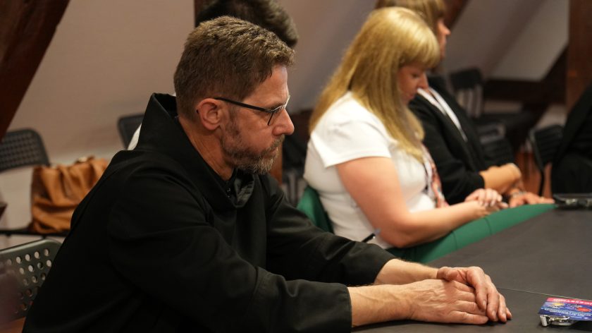 Brat zakonny w czarnym habicie siedzi przy stole konferencyjnym z rękami opartymi na blacie. Obok niego siedzą uczestnicy spotkania, a na stole widoczne są dokumenty i identyfikatory konferencyjne. Mężczyzna słucha wypowiedzi innych uczestników