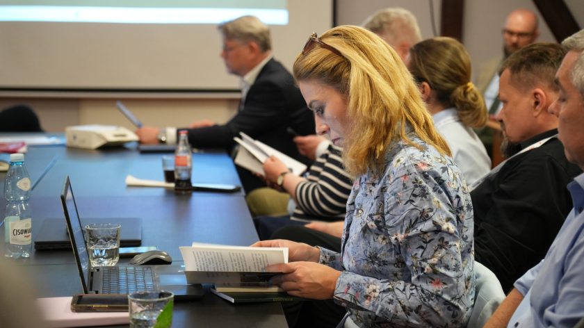 Kobieta w kwiecistej koszuli siedzi przy stole konferencyjnym i czyta książkę lub broszurę. Obok niej znajdują się laptop, szklanki oraz butelka wody mineralnej. W tle pozostali uczestnicy spotkania zajmują się materiałami konferencyjnymi