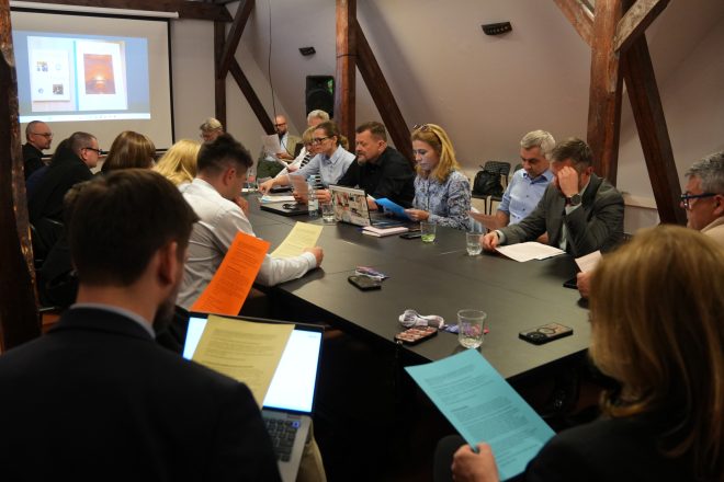 Szerokie ujęcie spotkania konferencyjnego z udziałem kilkunastu osób siedzących wokół dużego stołu. Uczestnicy czytają kolorowe materiały informacyjne i korzystają z laptopów. W tle widoczny jest ekran projekcyjny z prezentacją oraz drewniane belki poddasza