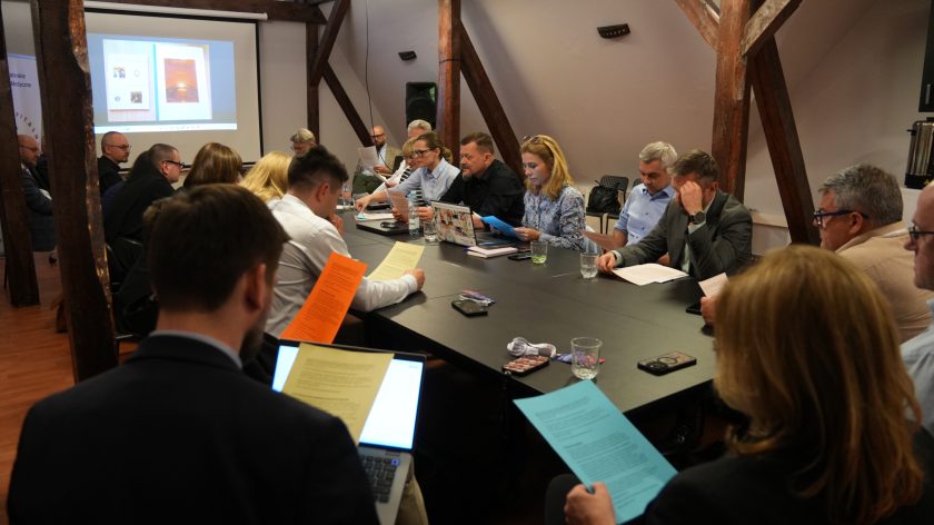 Szerokie ujęcie spotkania konferencyjnego z udziałem kilkunastu osób siedzących wokół dużego stołu. Uczestnicy czytają kolorowe materiały informacyjne i korzystają z laptopów. W tle widoczny jest ekran projekcyjny z prezentacją oraz drewniane belki poddasza