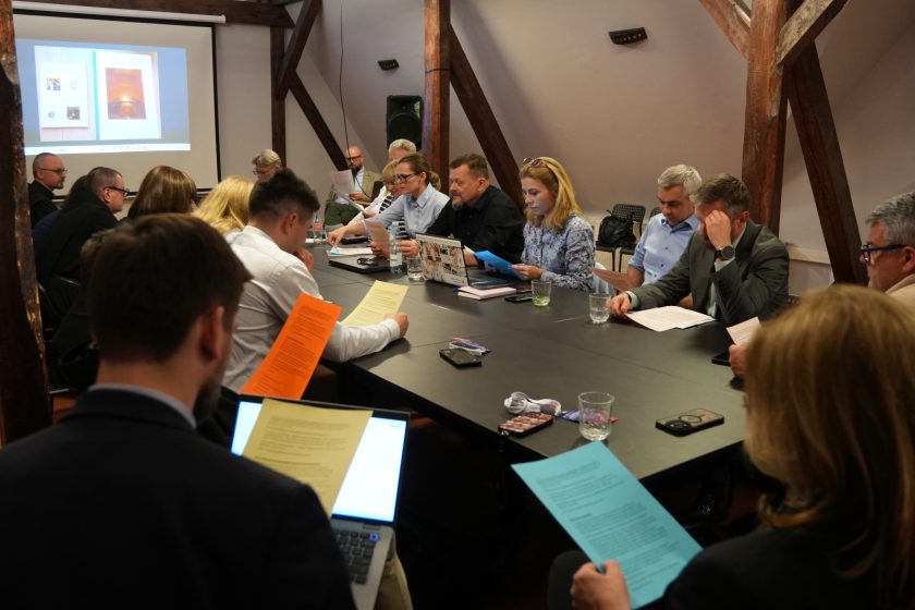 Szerokie ujęcie spotkania konferencyjnego z udziałem kilkunastu osób siedzących wokół dużego stołu. Uczestnicy czytają kolorowe materiały informacyjne i korzystają z laptopów. W tle widoczny jest ekran projekcyjny z prezentacją oraz drewniane belki poddasza