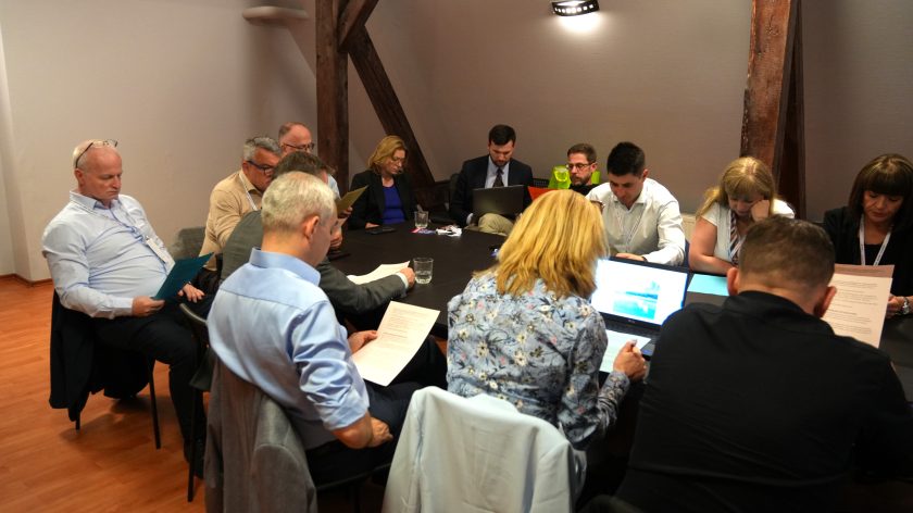 Uczestnicy spotkania siedzą przy stole konferencyjnym i analizują wydrukowane dokumenty. Na pierwszym planie kobieta pracuje przy laptopie z wyświetlonym wykresem, obok inni uczestnicy czytają materiały konferencyjne. Spotkanie odbywa się w sali na poddaszu z charakterystycznymi drewnianymi belkami